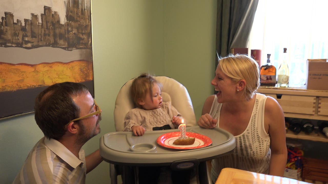 Toddler in a high chair with a lit candle as two adults lean nearby.