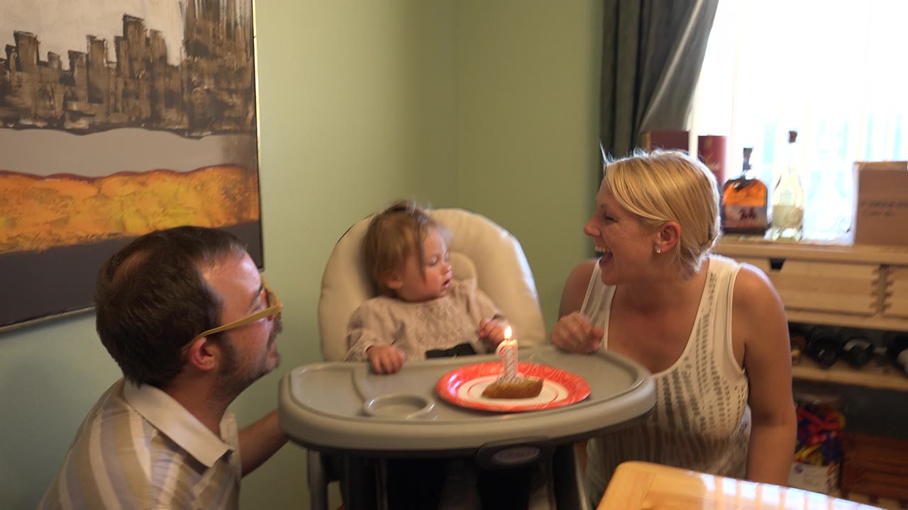 In Westminster, Colorado, the toddler looks down at the lit candle on a red plate as the woman smiles from the right and the man remains partly visible on the left.