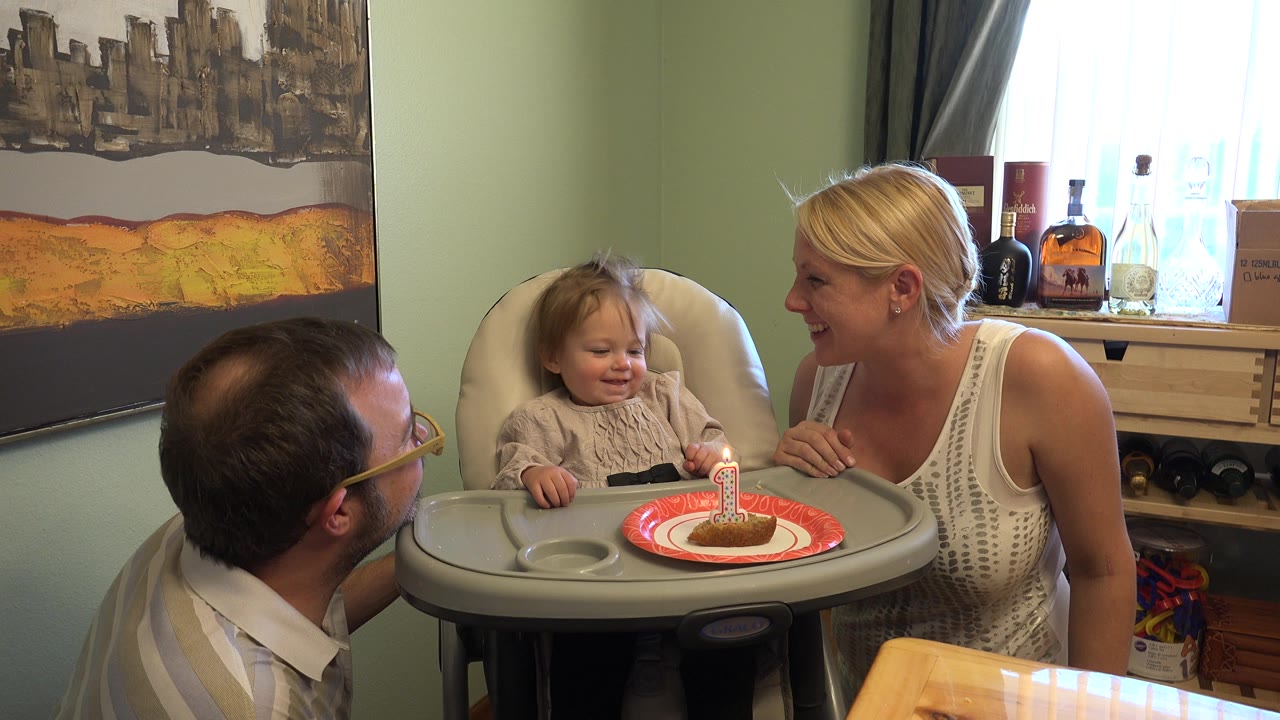Toddler in a high chair with adults leaning near a lit birthday candle.