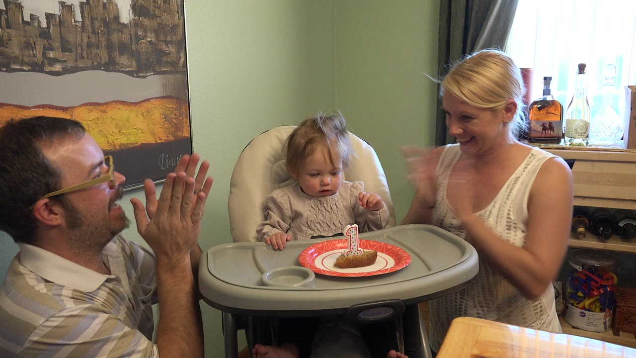 Man and woman clap beside a toddler in a white highchair with a small cake.