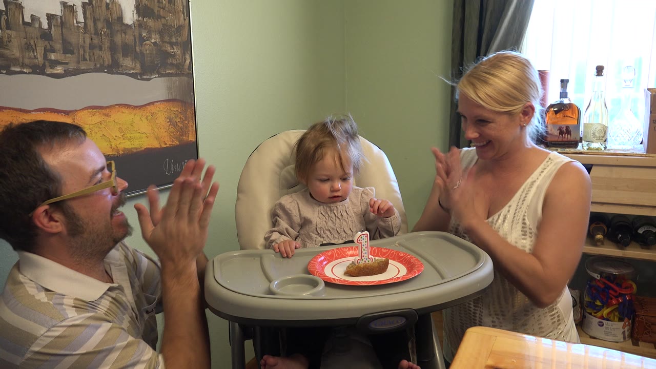 In Westminster, Colorado, the toddler looks toward the small cake on the highchair tray as the man and woman continue clapping nearby.