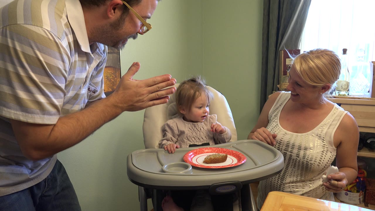 Father leans toward a child in a high chair while the mother sits nearby.