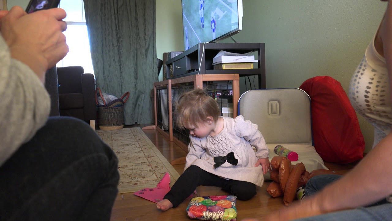 At the start of the Westminster, Colorado home clip, a toddler sits on the wood floor near a colorful gift bag while adult legs and a hand with a phone frame the foreground.