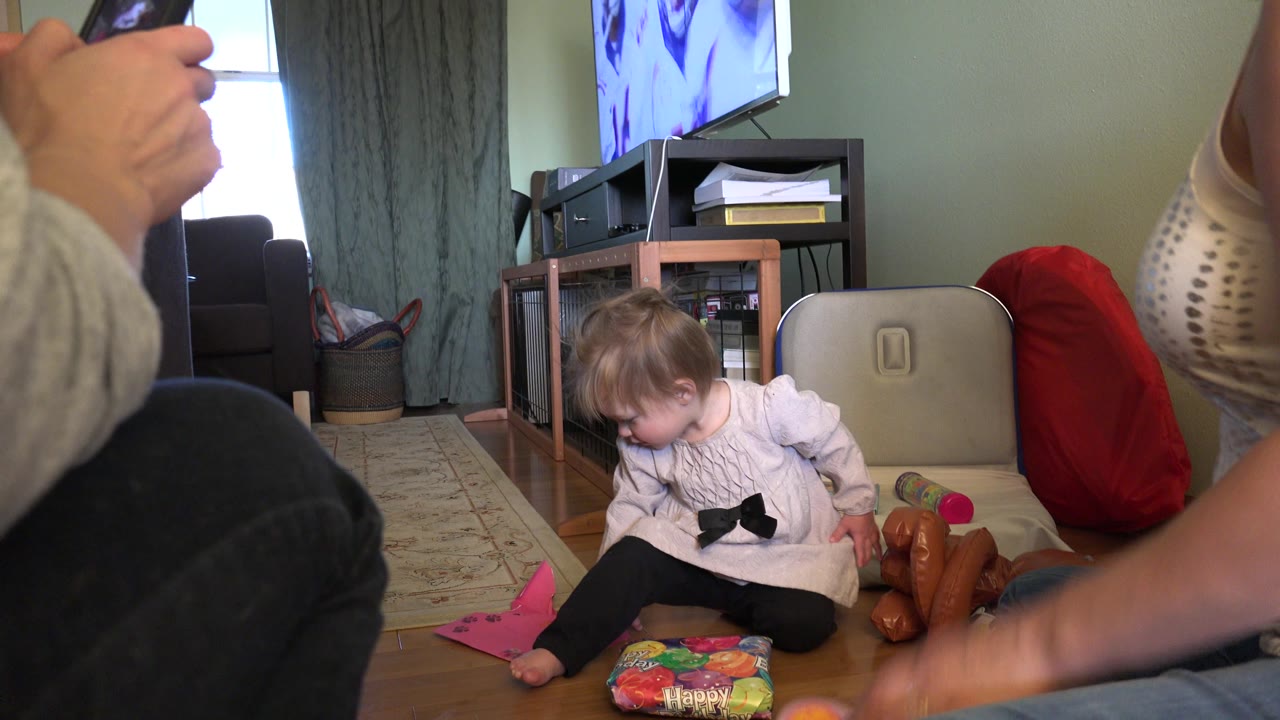 One second in, the toddler leans toward the gift bag on the living room floor as a seated adult remains close to the camera and the TV stand fills the background.