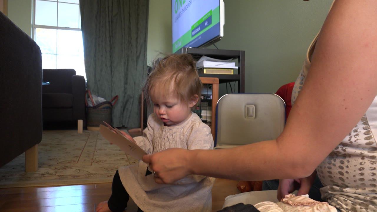 Blonde toddler holding a paper card beside an adult in a living room.