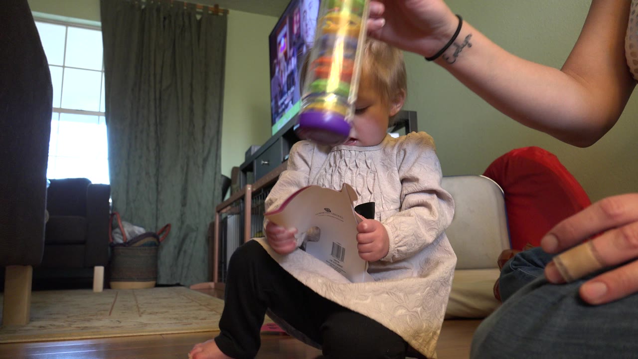 Baby seated on a living room floor as an adult holds a colorful tube toy overhead.