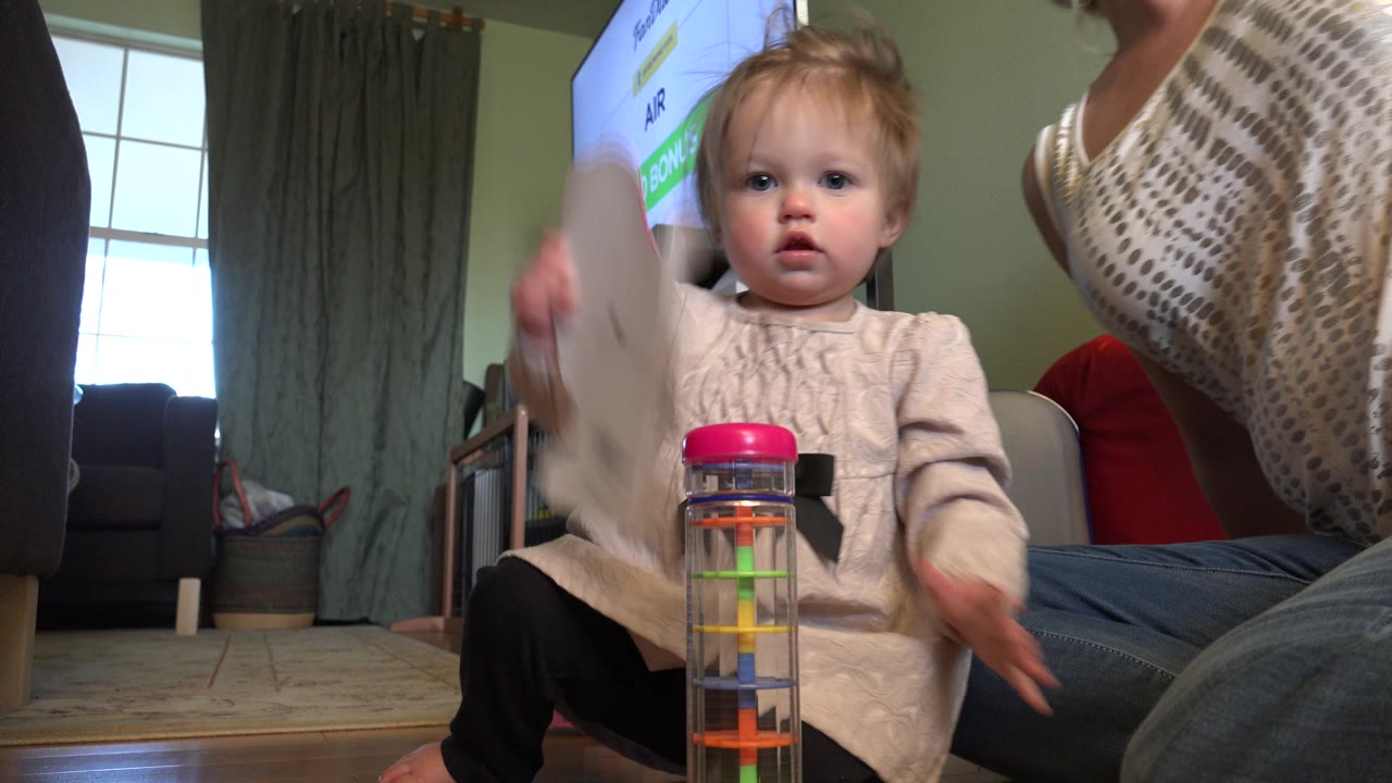 Young child on a living room floor beside a colorful stacking toy and adults.