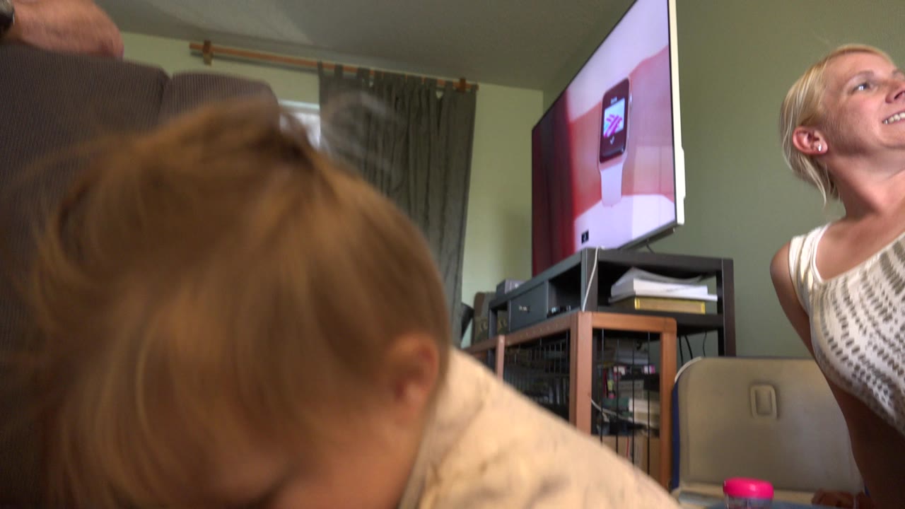 A baby fills the foreground near a floor-level camera in a Westminster, Colorado living room, with adult figures, a sofa, curtains, and a TV visible behind.