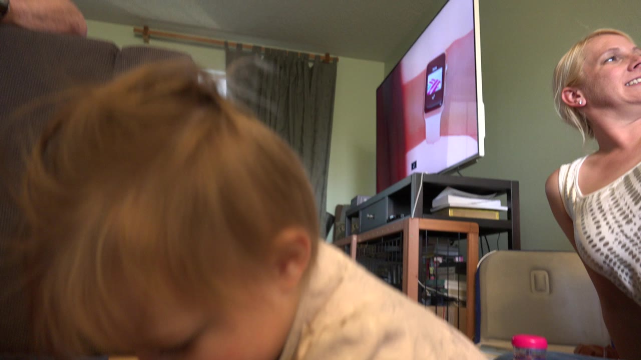 The baby remains close to the camera while a smiling young woman is visible near a television and media stand in the Westminster, Colorado home interior.