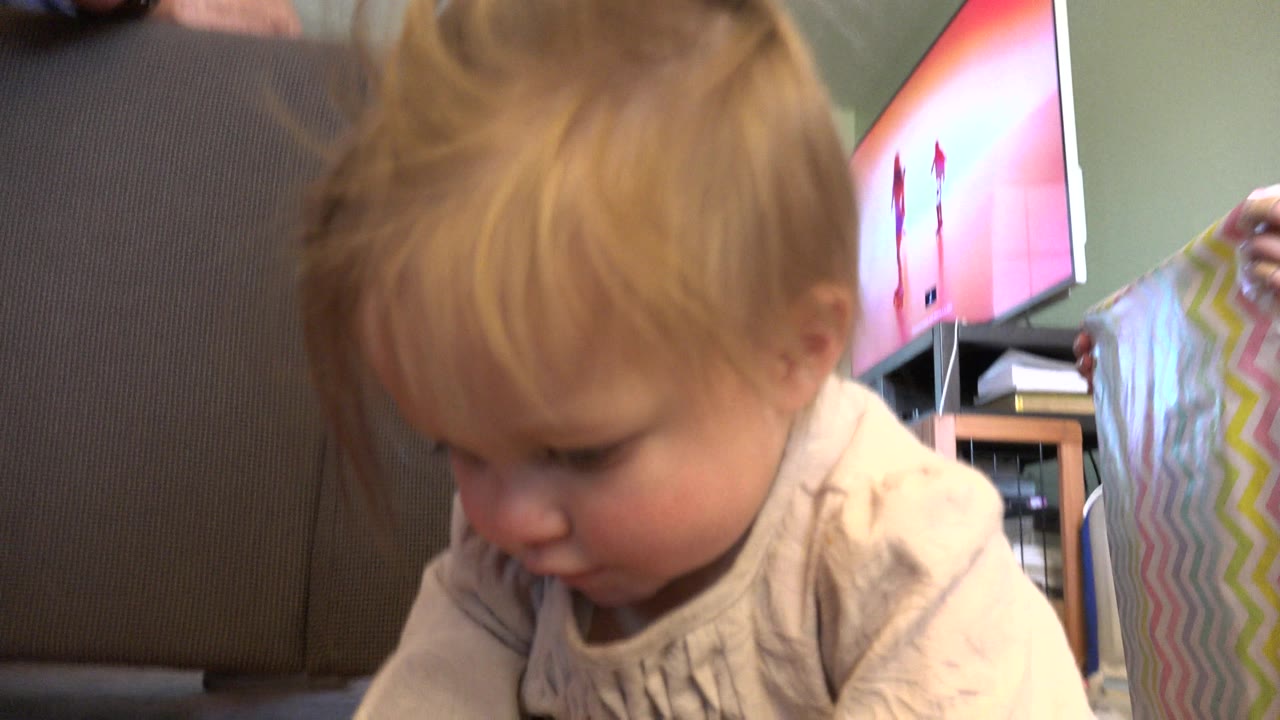 At 0:00 in Westminster, Colorado, a blond toddler in a light shirt looks down in close foreground beside a brown chair, with a pastel striped card held at the right edge.