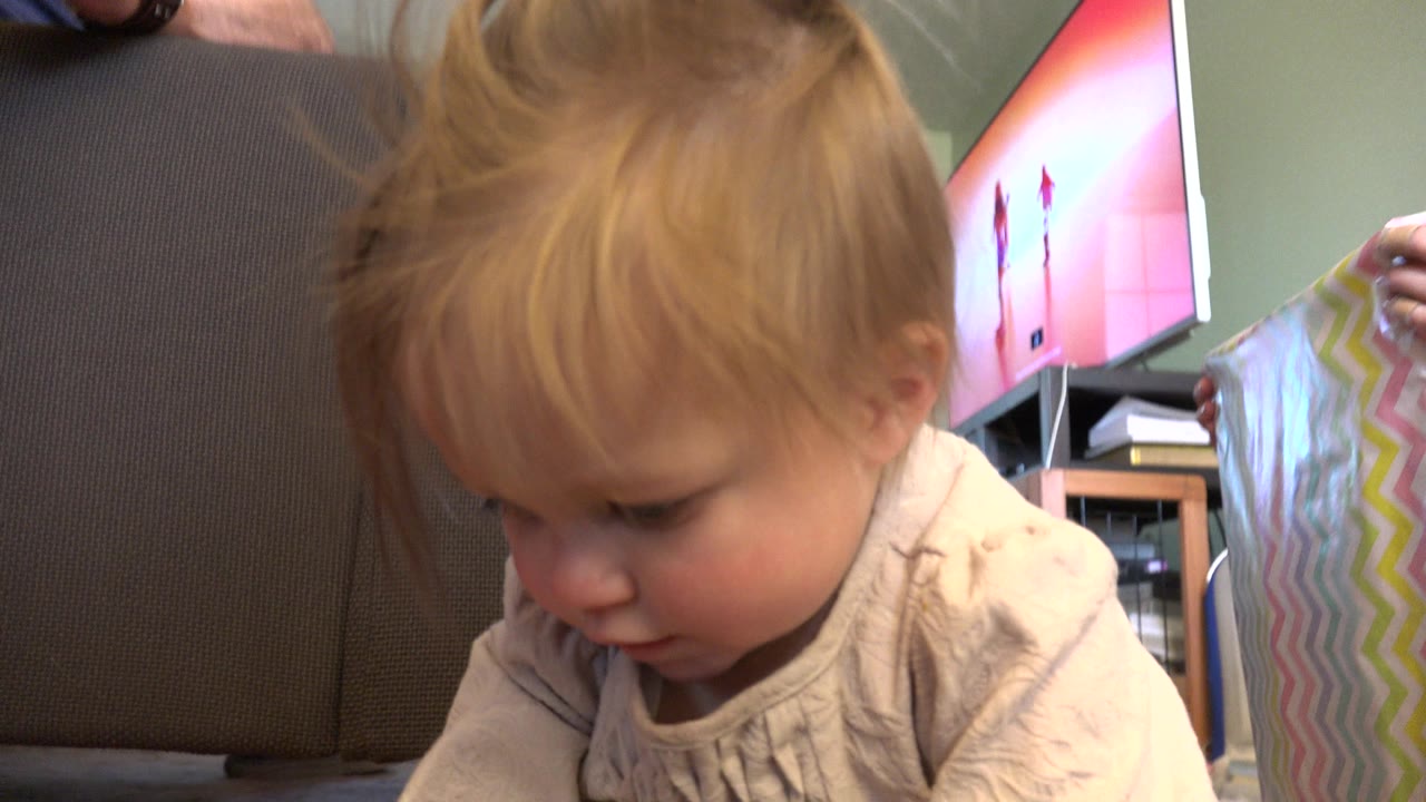At 0:01 in Westminster, Colorado, the toddler leans forward with eyes lowered while an adult hand holds the pastel striped card beside the brown chair.