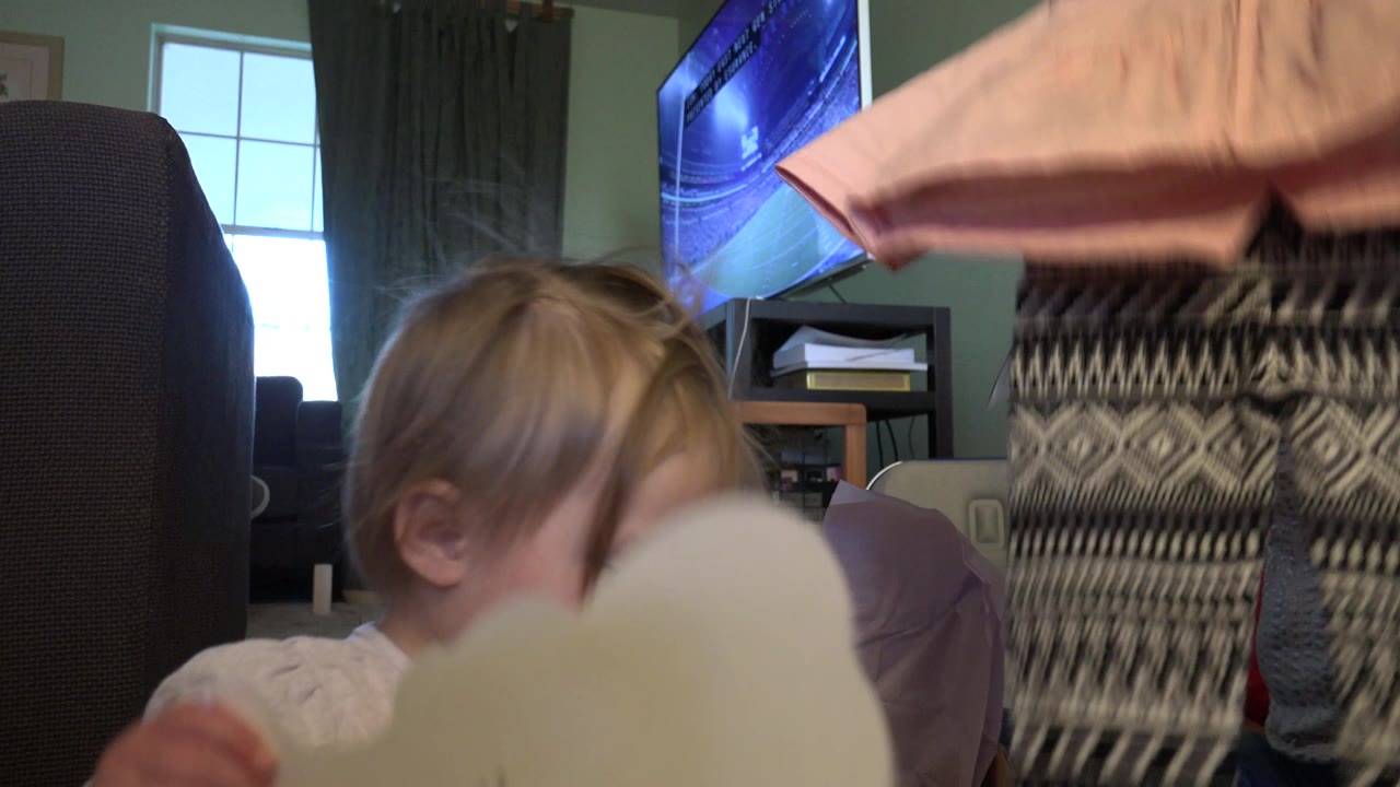 Toddler beside a pale card as clothing passes in front of the camera in a living room.
