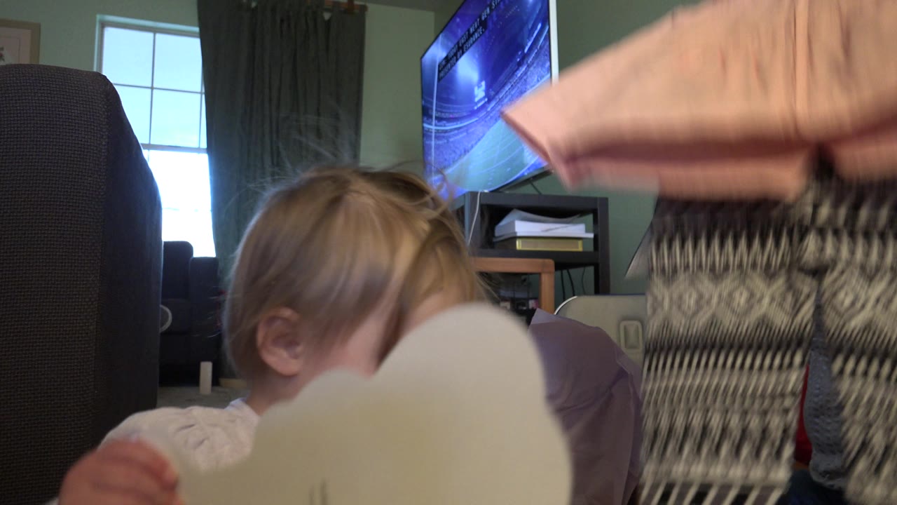 At 0:01 in Westminster, Colorado, the toddler remains close to the pale card while a pink clothing item and patterned leggings fill the center of the living room frame.
