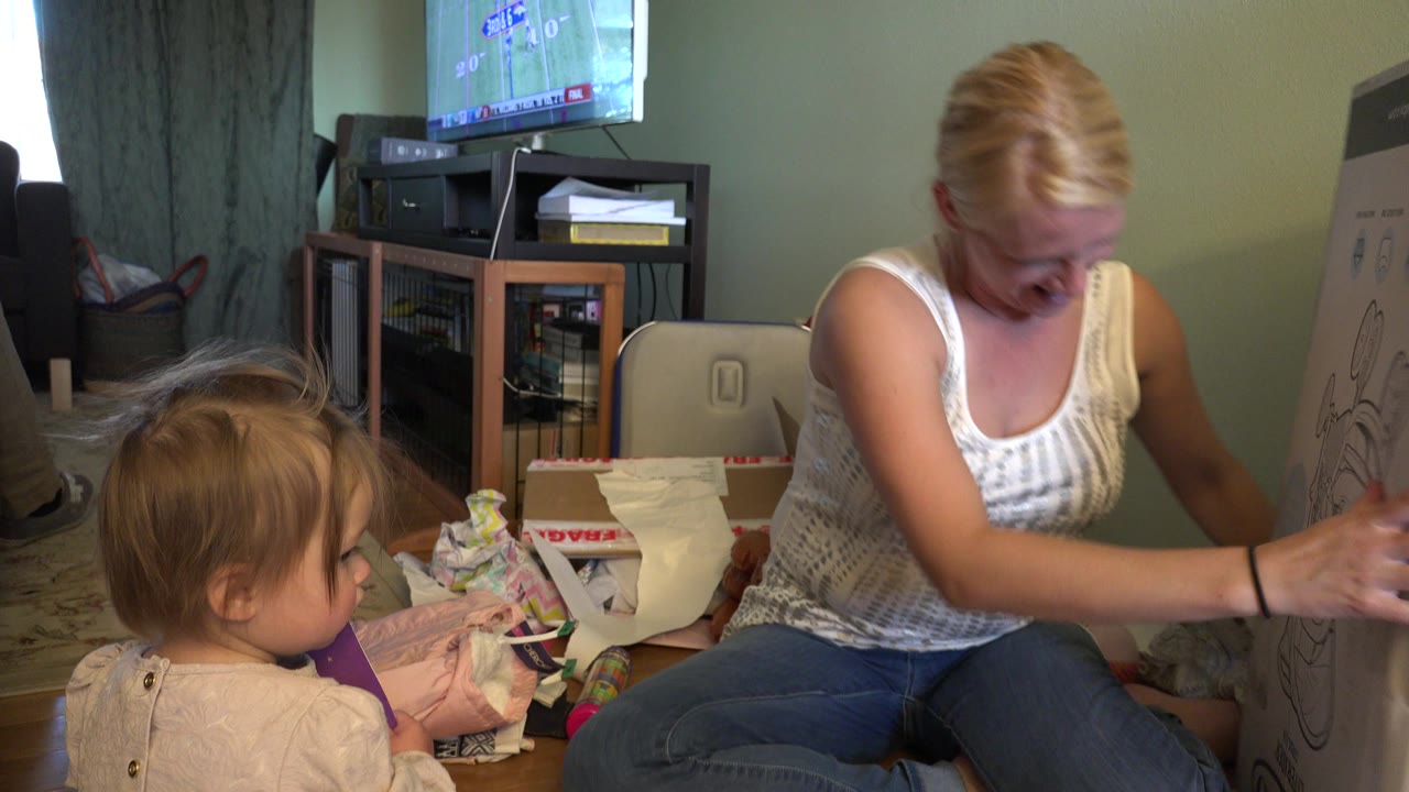 In Westminster, Colorado, a woman looks down while seated on the floor beside a toddler, with gift wrap, a TV, shelves, curtains, and a large illustrated box visible around them.