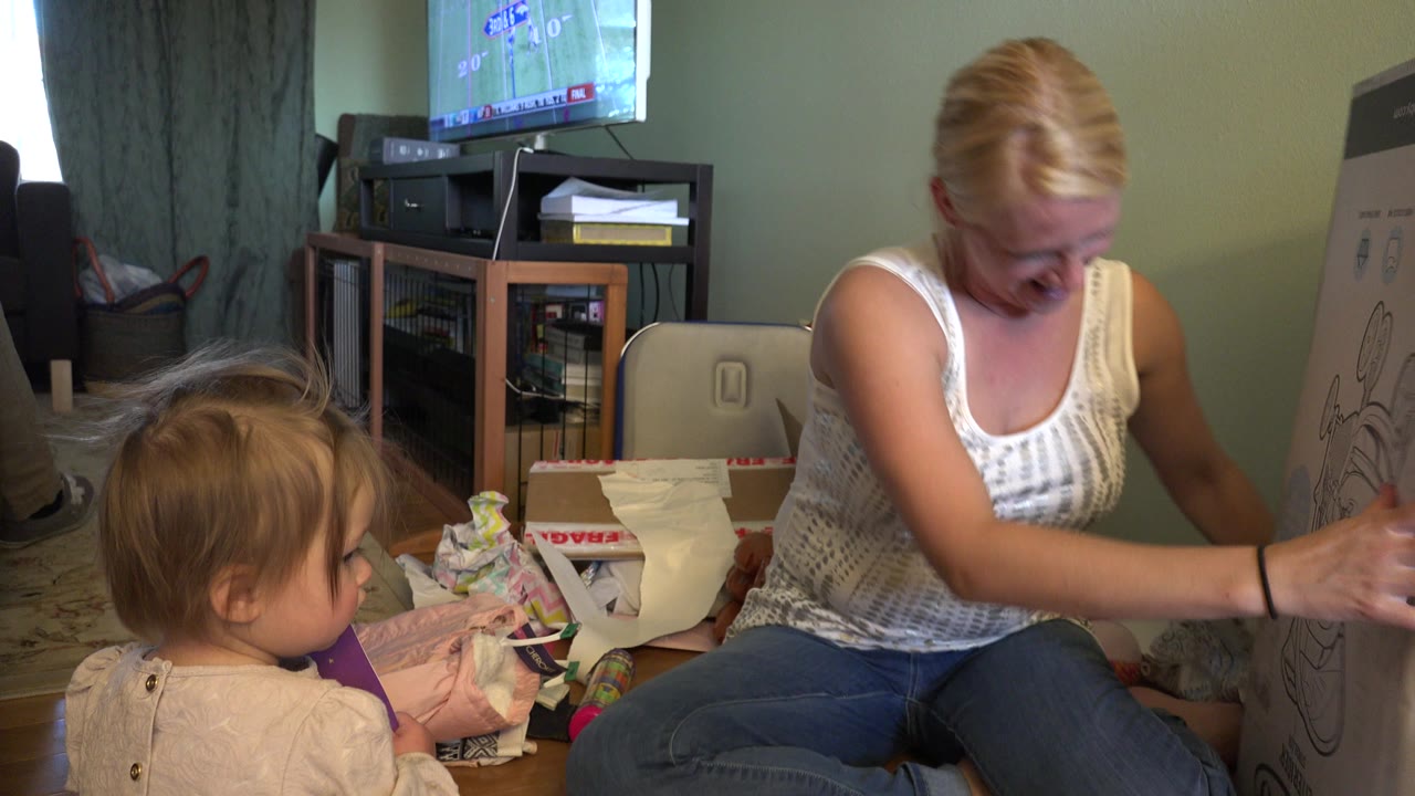 In Westminster, Colorado, the seated woman leans forward with her hand near the toddler, while the large illustrated box stands at the right edge of the living room frame.