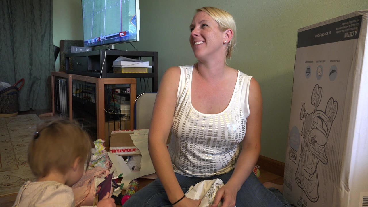 The toddler leans toward colorful gift paper in the foreground as the mother smiles near a TV stand and large boxed item in a Westminster, Colorado home.