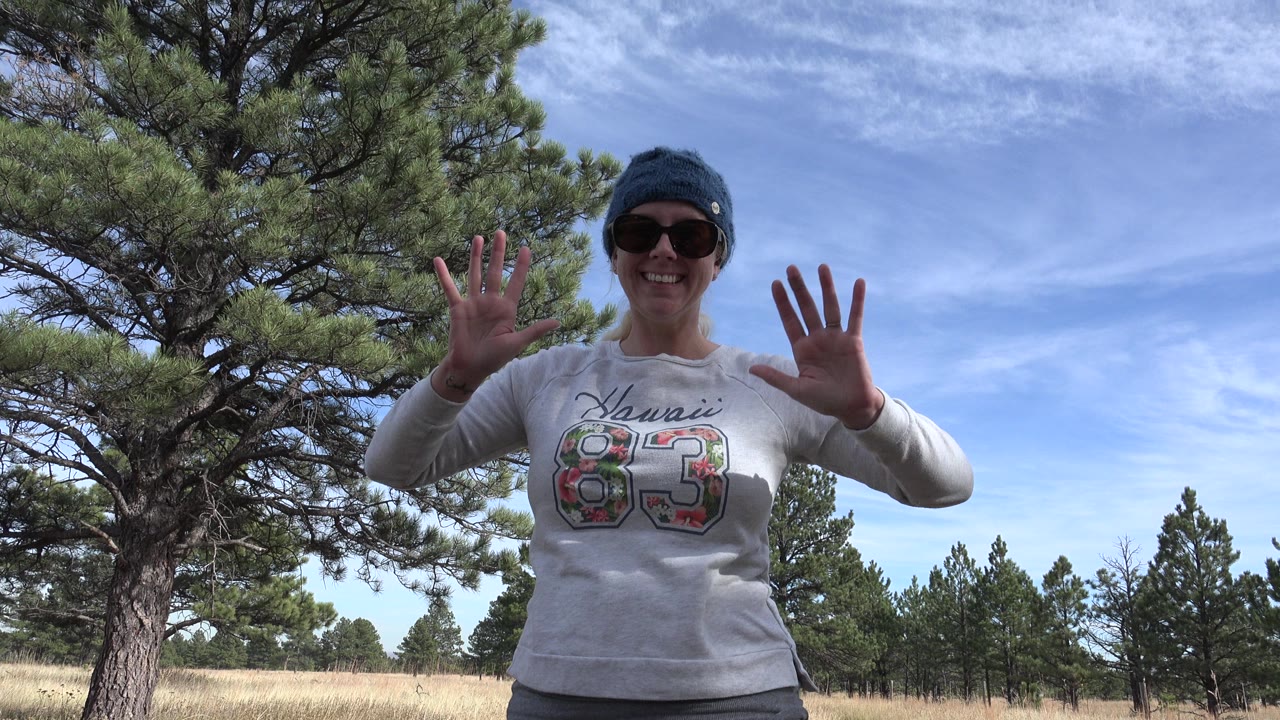 Woman in sunglasses waves on a grassy pine trail.