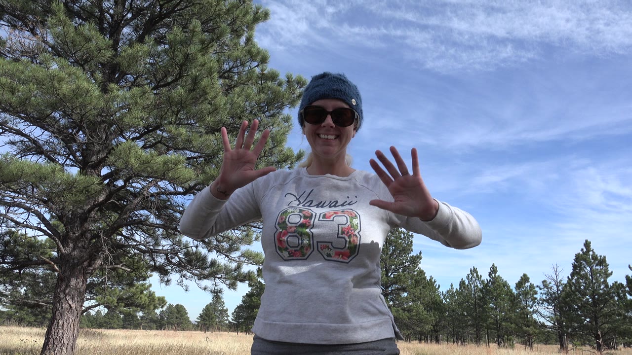 The woman continues waving with both hands raised in Golden, Colorado, framed from the waist up against pine branches, dry grass, and a partly cloudy blue sky.