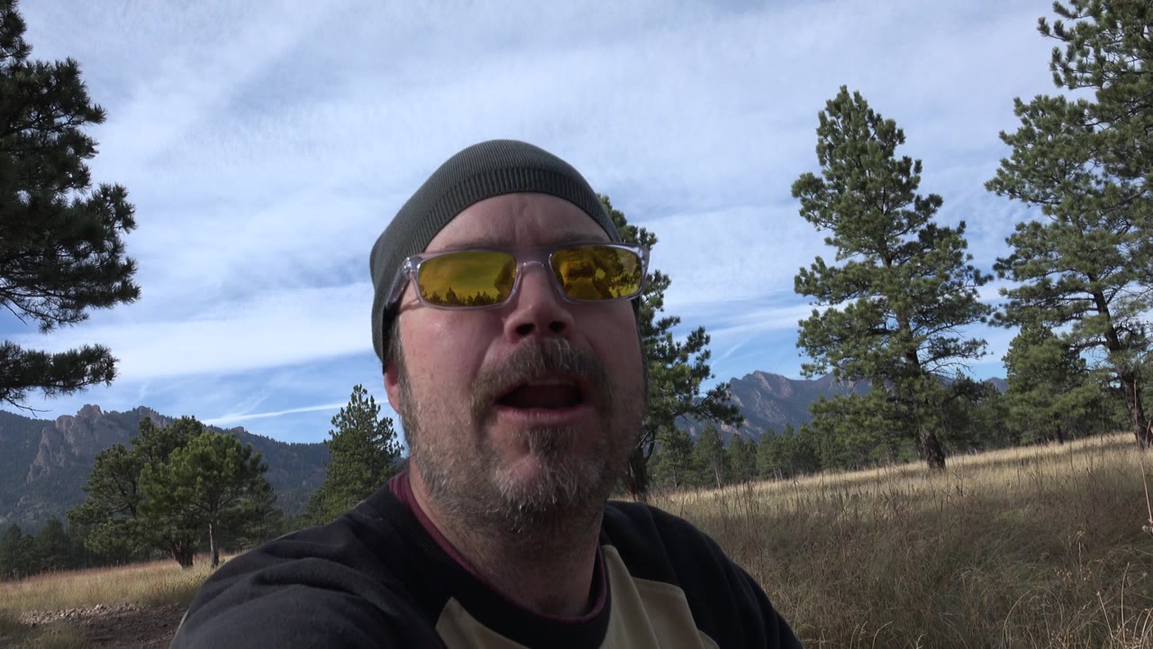 The man remains close in frame in Golden, Colorado, with yellow-tinted glasses reflecting the outdoor light and a pine meadow stretching behind his shoulder.