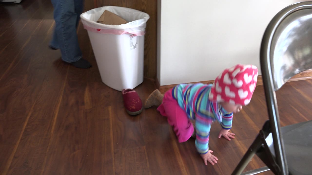 A baby in a pink heart hat crawls on a wood floor in a Prairie Grove, Illinois home, with a gray folding chair in the foreground and a white bin by the doorway.