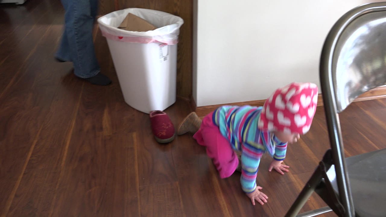 The baby continues crawling across the wood floor toward the gray folding chair in the Prairie Grove interior, with shoes and a white bin visible behind.
