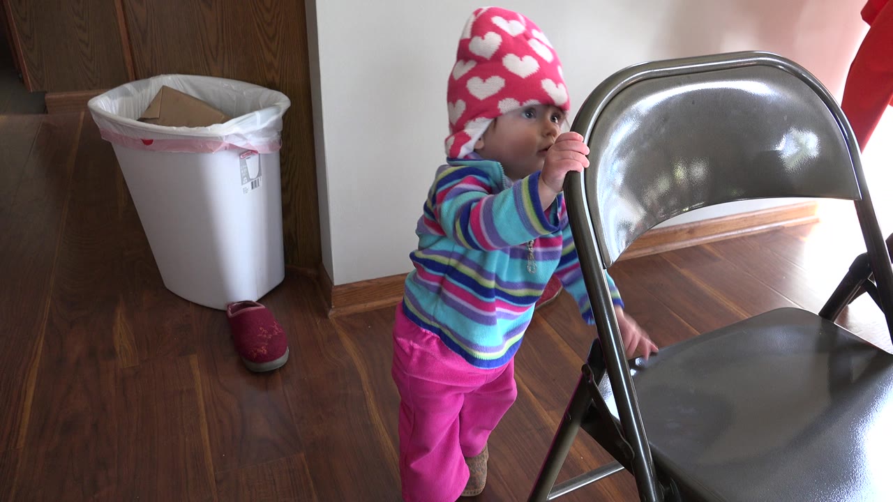 A baby in a pink heart hat and striped shirt stands beside a metal folding chair in a Prairie Grove, Illinois home interior, with a trash bin and wood floor visible.