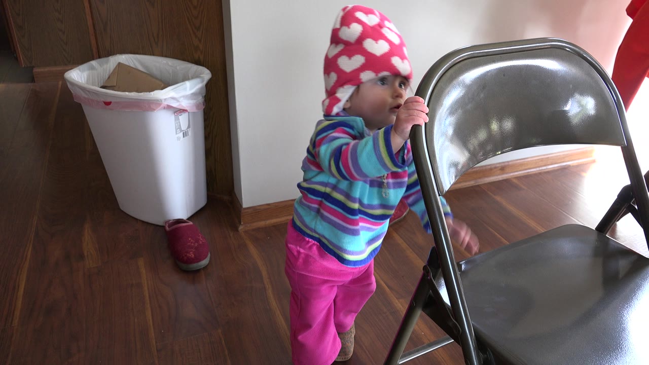 The baby holds the chair back while looking toward the room inside a Prairie Grove, Illinois home, with the white trash bin and doorway still in the background.
