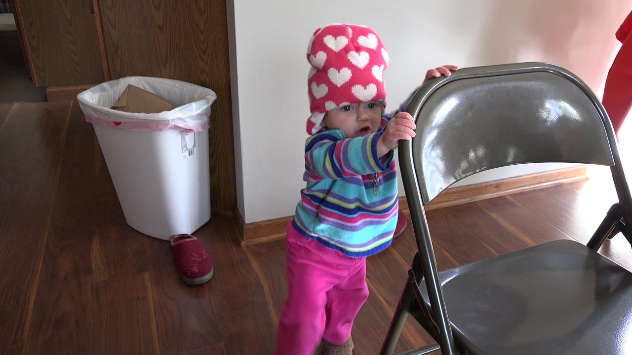Baby partly behind a metal folding chair with one hand on the chair back.