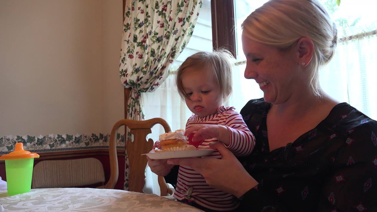 Toddler on a woman's lap reaching toward a cupcake on a plate.