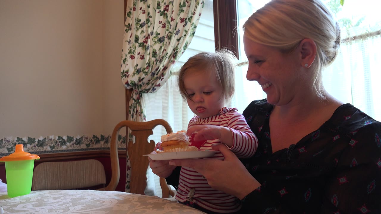 The toddler in a red striped shirt touches the cupcake as the blonde woman smiles, with floral curtains and soft daylight behind them in Prairie Grove, Illinois.
