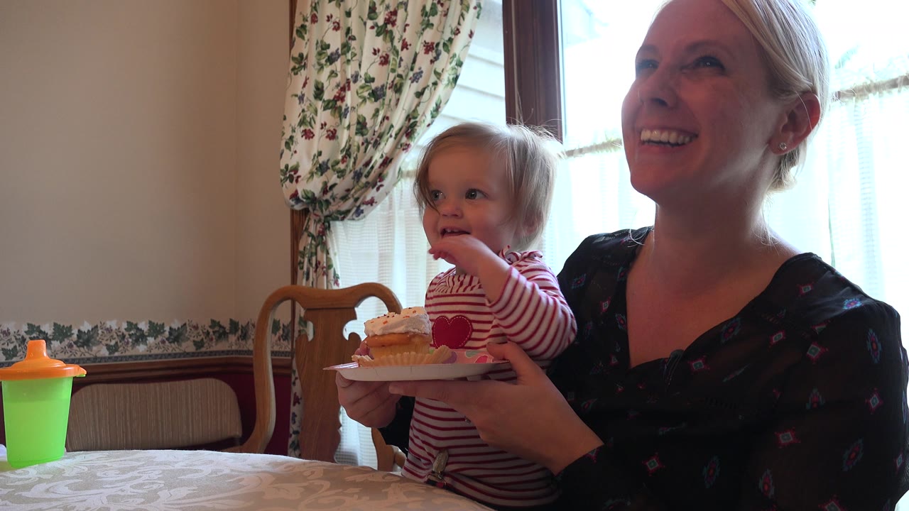 Toddler with cupcake held by smiling woman at a dining table.