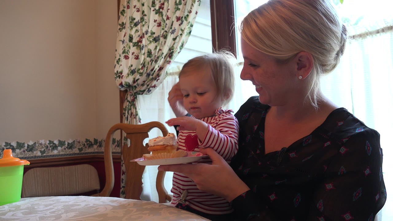 In Prairie Grove, Illinois, a toddler sits on a mother's lap and reaches toward a cream-topped cupcake on a plate near a bright dining room window.