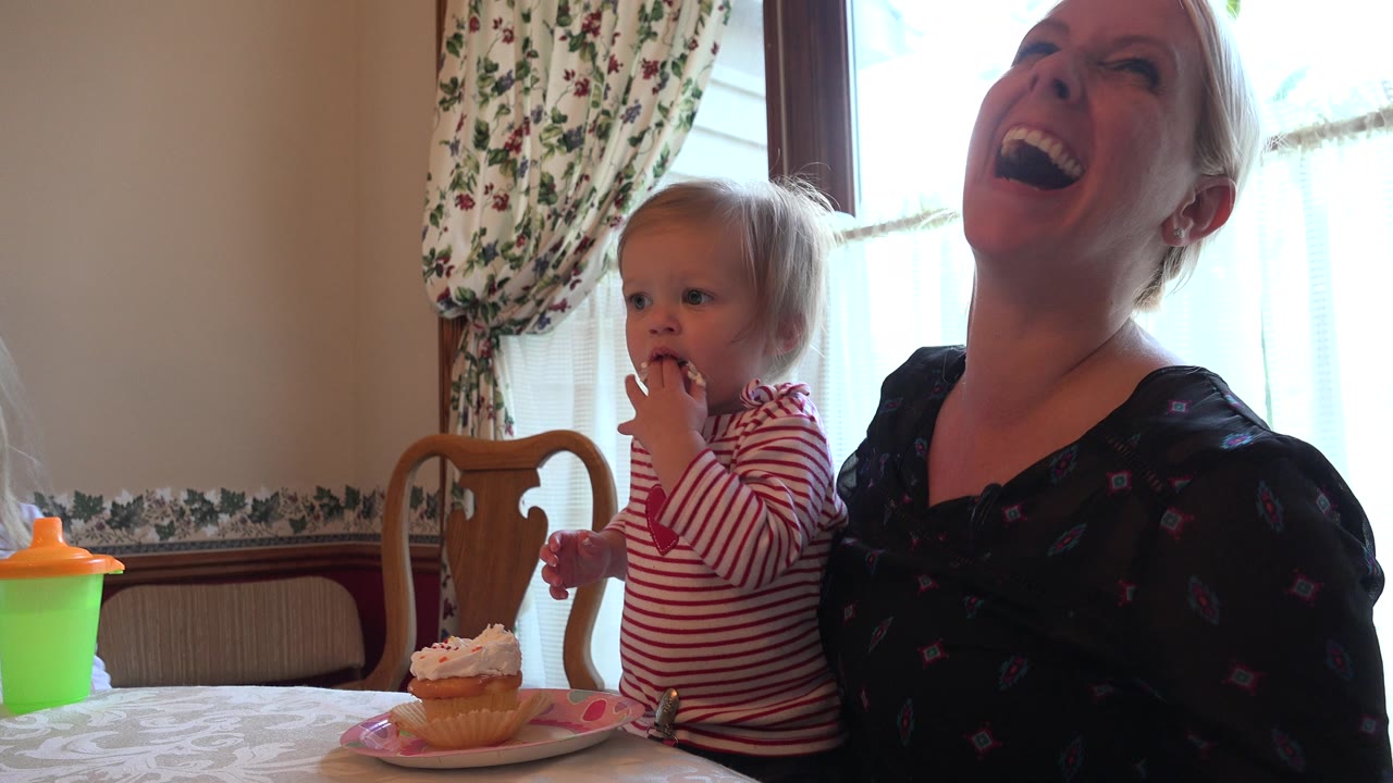 In Prairie Grove, Illinois, a toddler in a red striped shirt tastes frosting from a cupcake while her mother laughs beside her near a bright window.