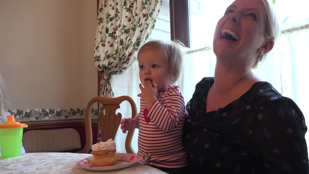 In Prairie Grove, Illinois, the toddler keeps her fingers near her mouth while the cupcake sits on the plate and her mother laughs in the right side of the frame.