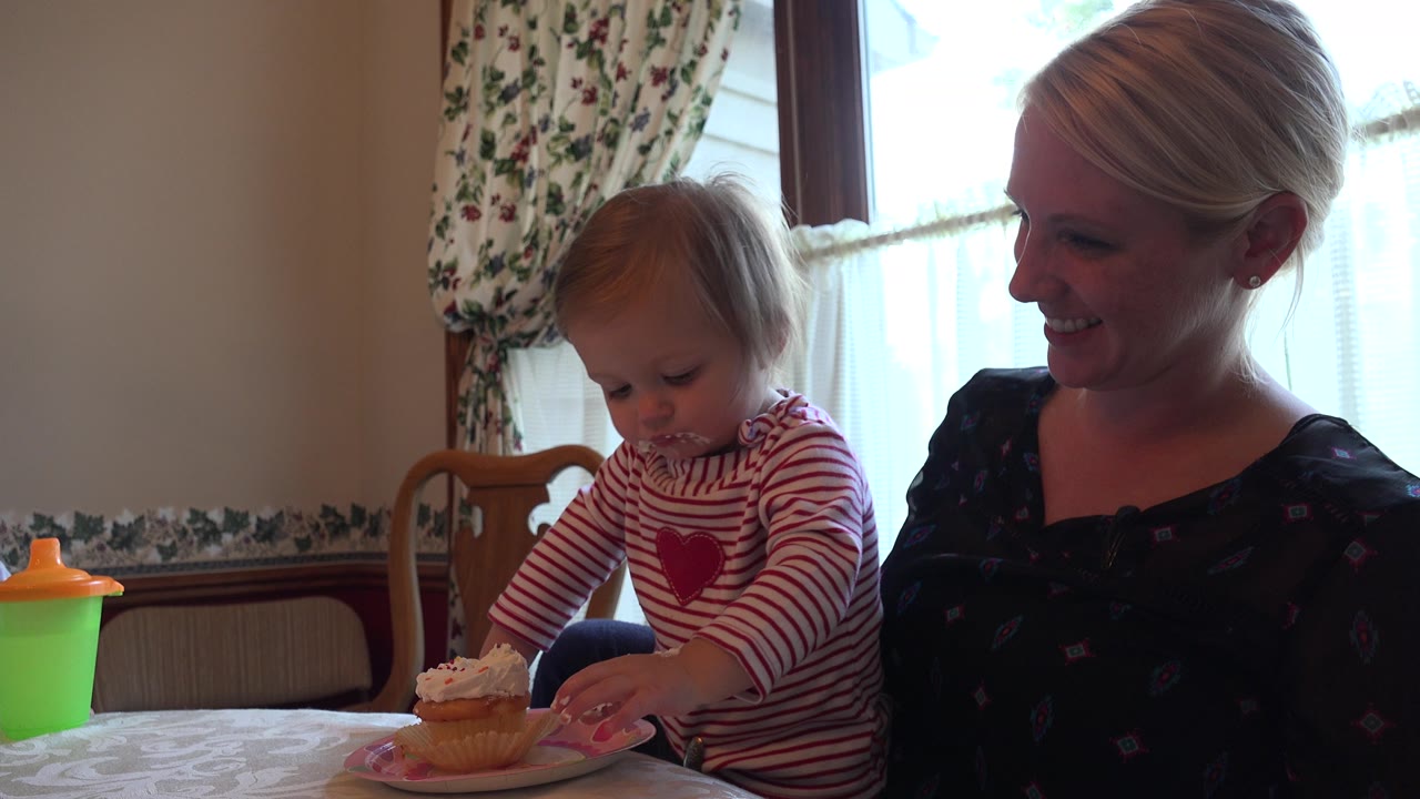 Toddler in a red striped shirt stands beside a cupcake while a woman smiles nearby.