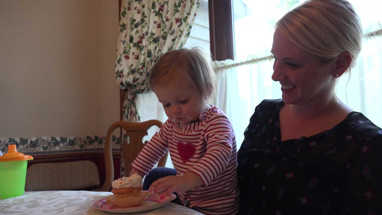 In Prairie Grove, Illinois, the toddler looks down at the cupcake plate while the seated woman smiles, with window light behind them and a green sippy cup at the table edge.
