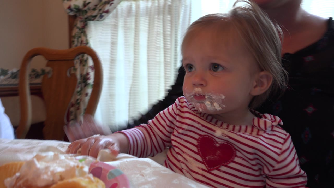 A toddler with cream around their mouth sits at a dining table in Prairie Grove, Illinois, with an adult partly visible behind and sheer curtains in the background.