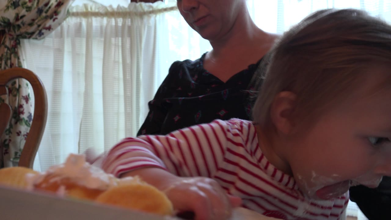 Toddler leans toward food at a dining table with an adult behind her.