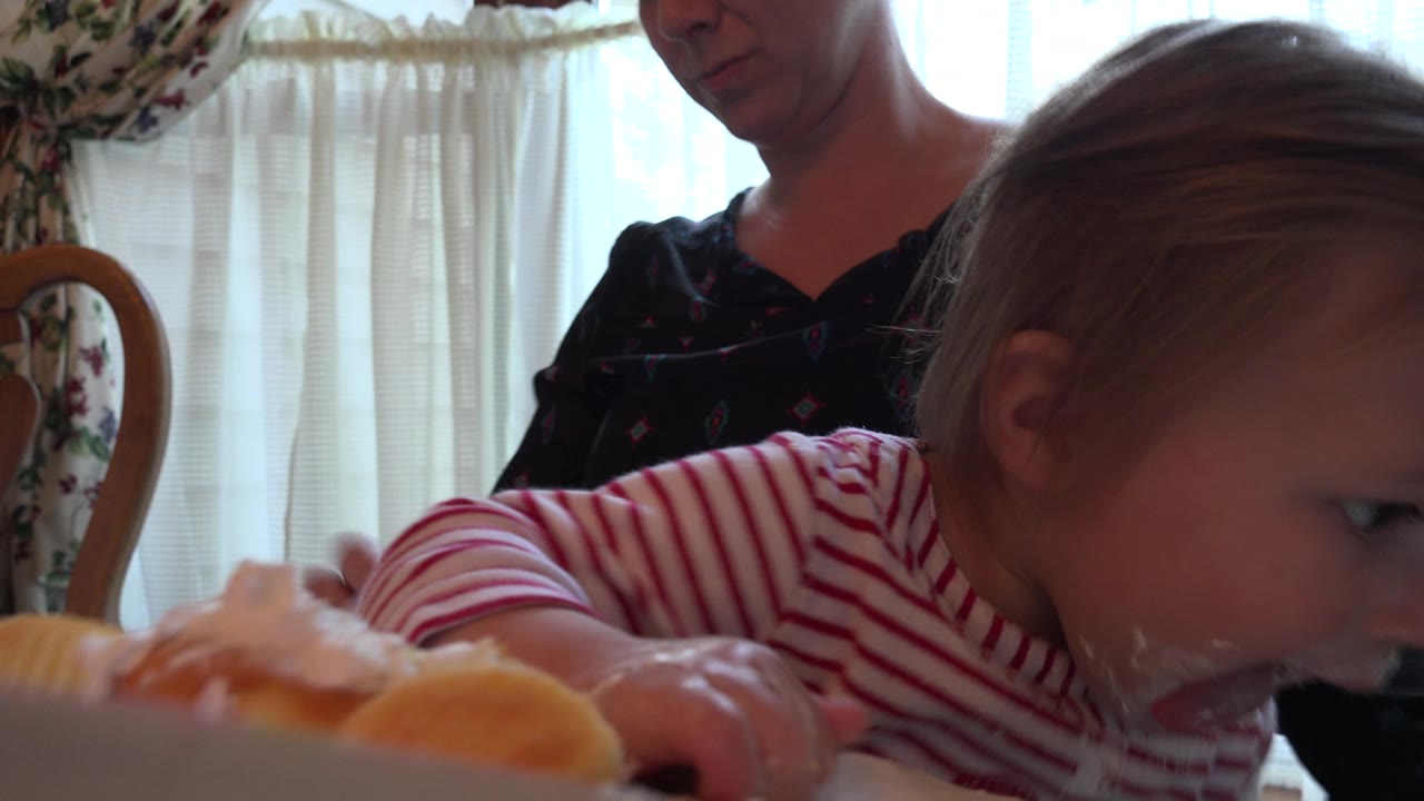 In Prairie Grove, Illinois, the toddler stays bent toward the foreground food, with the adult partly visible behind her in a softly backlit dining room.