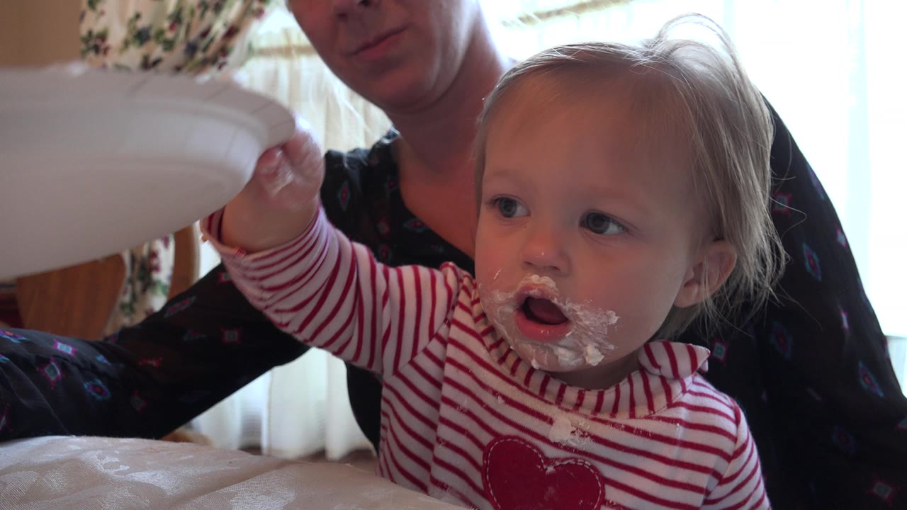 In Prairie Grove, Illinois, a young child in a red striped shirt reaches toward a white plate while an adult is partly visible behind in a bright dining room.