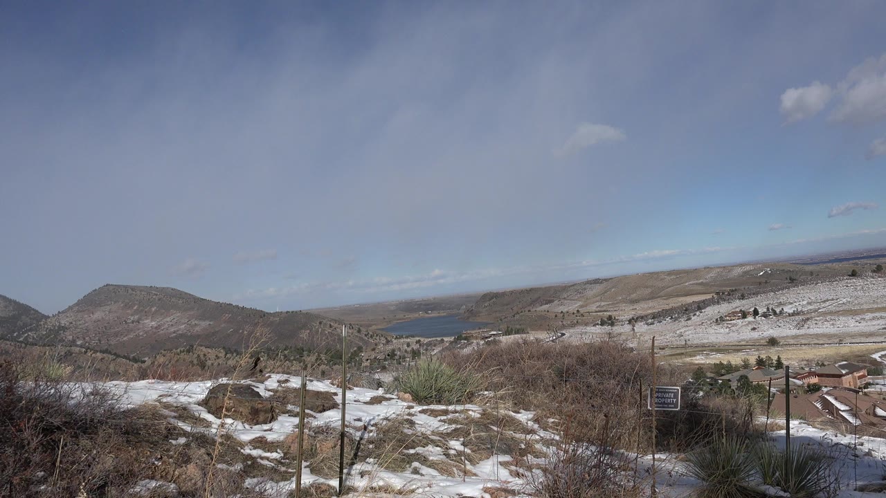 Snowy hillside overlook with brush, mountains, distant water, and blue sky.