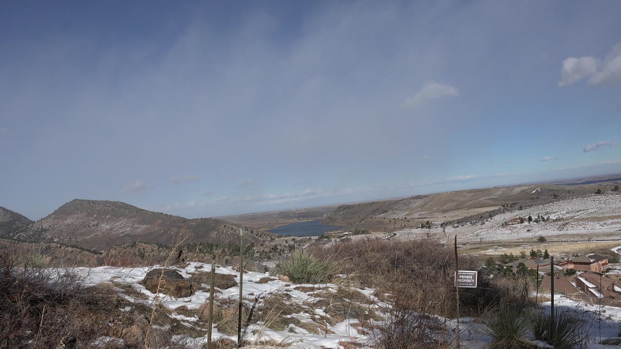Snowy dry hillside with brush, fence posts, distant hills, and blue sky.