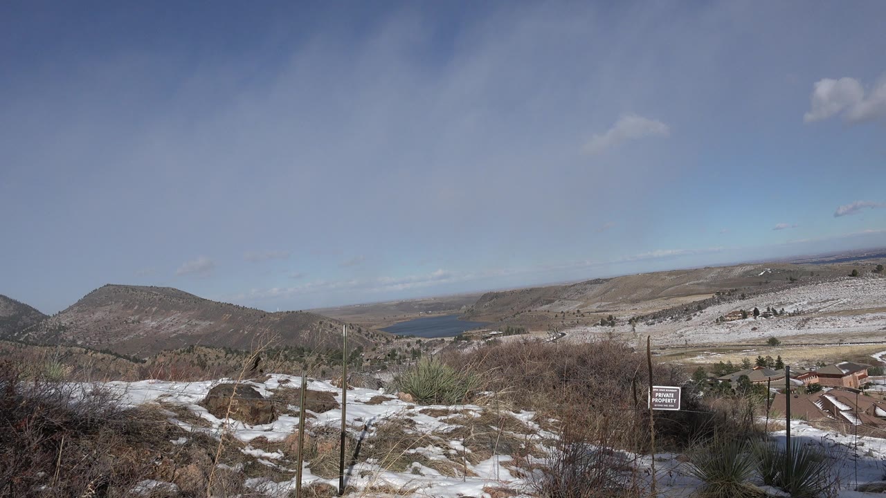 Snow-patched hillside with brush, fence posts, distant hills, and blue sky.