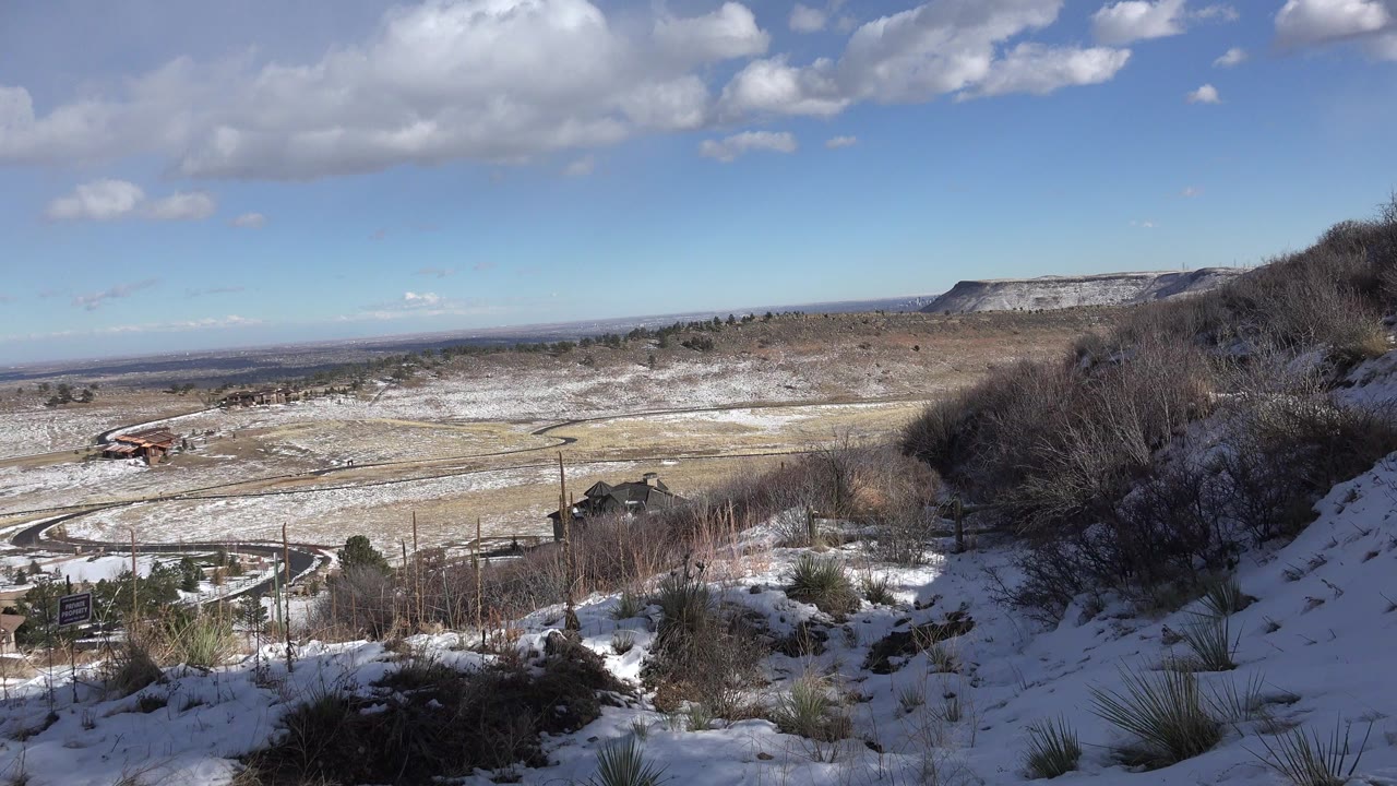Opening view in Golden, Colorado of a snowy hillside foreground, dry brush, a valley with curved roads, distant ridges, scattered clouds, and no visible people.