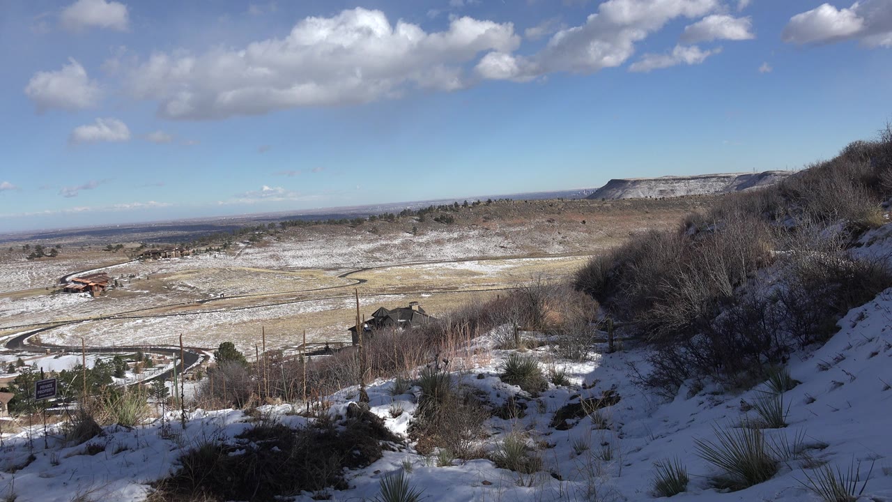 Snowy hillside overlook with open land, road, distant mesa, and blue sky.