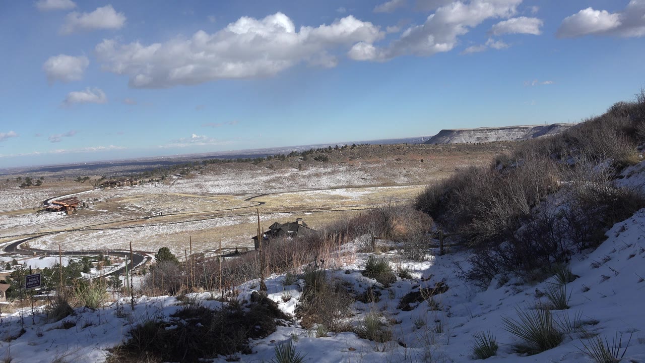 Snow-dusted overlook with dry brush, valley, roads, ridge, and blue sky.