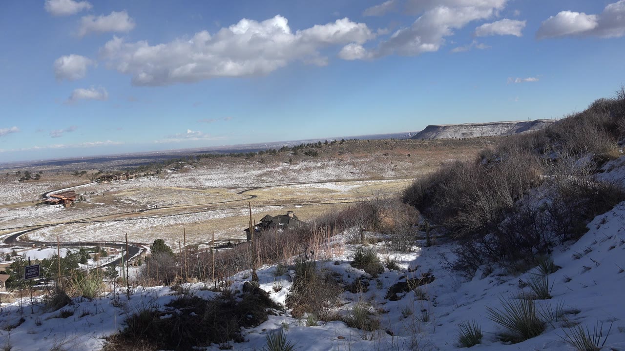 Opening frame of a Golden, Colorado hillside view with snow on grass, bare shrubs at right, a curving road in the valley below, scattered clouds, and no visible people.