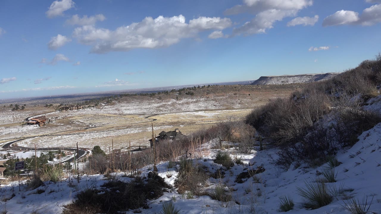 Snowy hillside overlook with valley roads, scattered buildings, ridge, and blue sky.