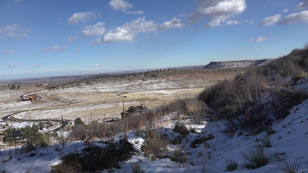 Opening view in Golden, Colorado from a snowy hillside with brush in front, roads and scattered buildings below, distant mesas, blue sky, and no visible people.