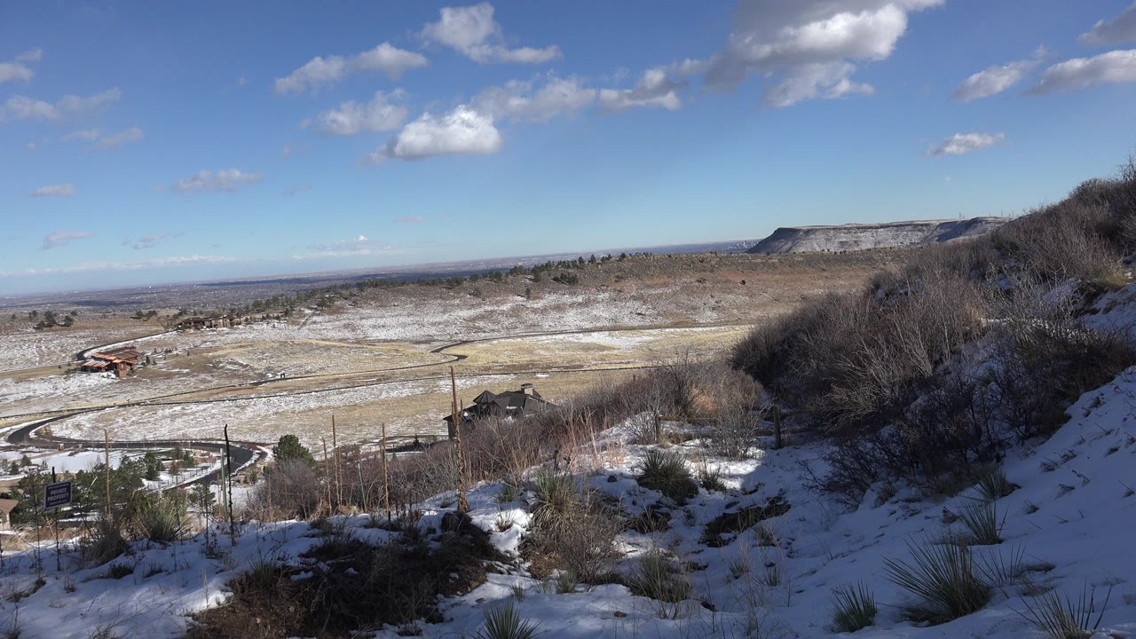 Opening view from a snowy hillside in Golden, Colorado, looking over roads, scattered buildings, a distant ridge, and blue sky with small clouds, with no visible people.